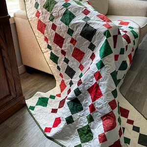 Puede incluir: Una colcha blanca con un patrón de diamantes en rojo y verde. La colcha está doblada y drapeada sobre un sofá.