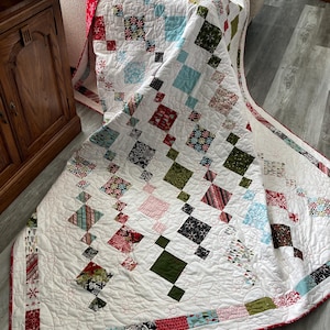 May include: A white quilt with a patchwork design featuring red, green, and blue fabrics. The quilt is folded and draped over a couch.