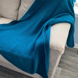 May include: A teal blue crocheted throw blanket draped over a light beige couch. The couch is in a room with wood-look flooring.