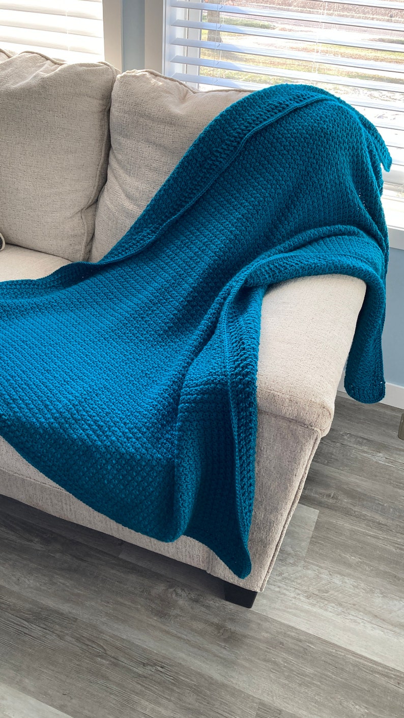May include: A teal blue crocheted throw blanket draped over a light beige couch. The couch is in a room with wood-look flooring.