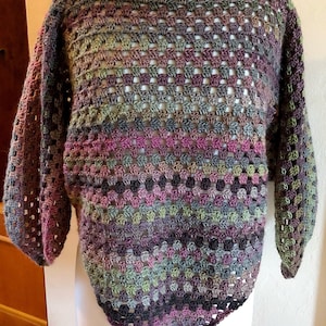 May include: A crocheted top with a triangular shape and a loose fit. The top is made with a multi-colored yarn in shades of green, purple, and brown.