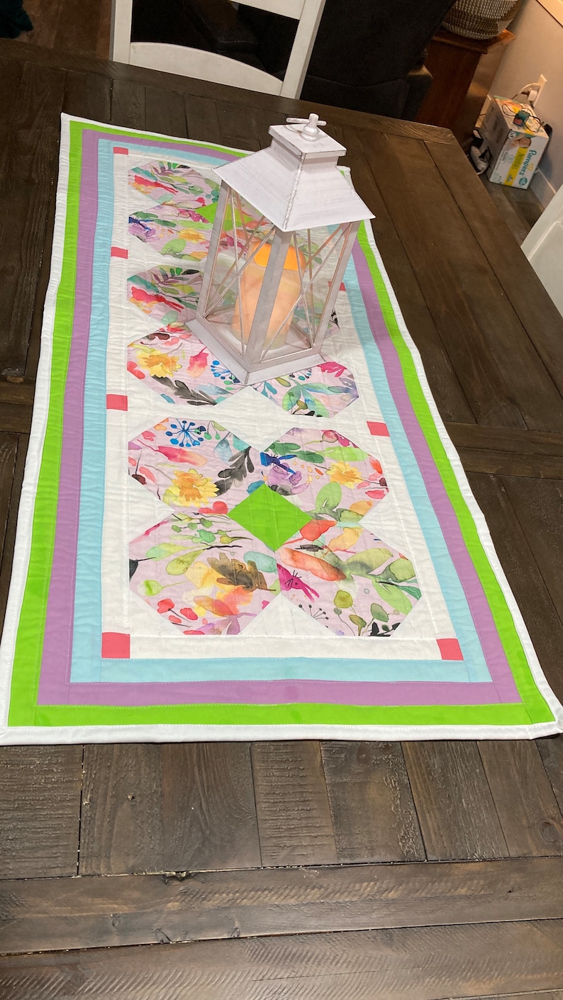 May include: A white table runner with a green, purple, and blue border. The runner has a floral pattern in the center and a white lantern on top of it. Beginner-Friendly Table Runner Pattern, Whimsical Springtime Bunnies Table Runner Quilt Pattern, Spring table runner pattern