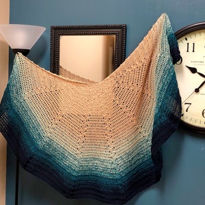 Puede incluir: Un chal tejido a crochet con un degradado de colores turquesa, azul y beige. El chal está drapeado sobre un espejo y un reloj.