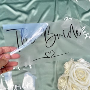 May include: A transparent sheet with the words "The Bride" in black script, and a heart symbol. The sheet is held by a hand, with a bouquet of white roses in the background. The background is a pale green.