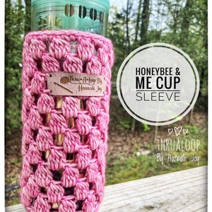 Puede incluir: Una funda de ganchillo rosa envuelta alrededor de un vaso de plástico transparente con tapa verde claro. Una etiqueta de cuero rectangular está unida al frente con el texto Thru-A-Loop By Hannah Joy. El texto Honeybee & Me Cup Sleeve es visible.