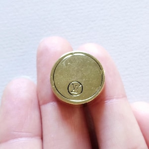 Puede incluir: Un botón redondo de color dorado con un logotipo LV negro en relieve en el centro.