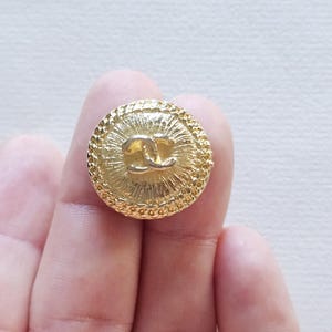 Peut inclure: Un bouton rond doré présentant un motif texturé en rayons de soleil partant du centre. Le logo emblématique CC entrelacé est gravé au milieu, entouré d'une bordure perlée décorative le long du bord extérieur du bouton circulaire.
