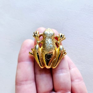 Op de afbeelding: Een gouden broche in de vorm van een kikker met een gestippeld patroon. De kikker zit op zijn achterpoten met zijn voorpoten omhoog.