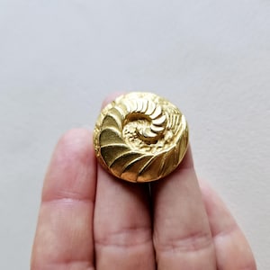 May include: A gold-toned metal button with a spiral design. The button has a raised, textured surface.