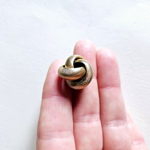 Puede incluir: Un colgante de metal dorado en forma de nudo. El colgante es texturizado y tiene un acabado liso y pulido.