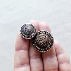 May include: Two antique-style, round buttons with a detailed face design. The buttons are brown with a silver-coloured border. One button is smaller than the other. The buttons are likely for clothing or craft projects.
