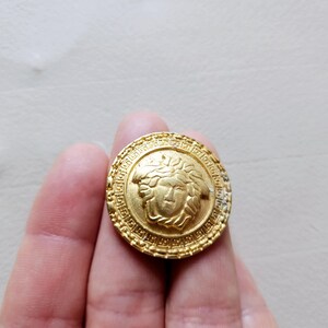 Könnte beinhalten: Goldfarbener Metallknopf mit einem erhabenen Design eines Gesichts mit einer Krone. Der Knopf hat einen detaillierten Rand.
