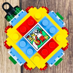 May include: A colorful, geometric ornament constructed from LEGO bricks. The design features red, yellow, blue, and green blocks, with a central square image of a raccoon. The ornament has a black loop for hanging.