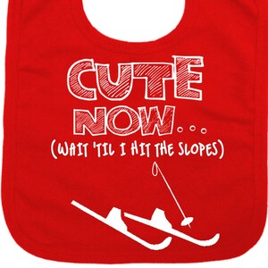 May include: Red baby bib with white text that says "Cute Now... (Wait 'til I hit the slopes)" and a white illustration of a skier.