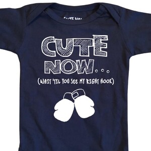 May include: Navy blue baby onesie with the text "CUTE NOW... (WAIT 'TIL YOU SEE MY RIGHT HOOK)" in white. The onesie features a pair of white boxing gloves below the text. The onesie has short sleeves and snaps at the bottom.