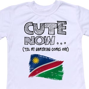 Peut inclure: T-shirt blanc avec un texte noir qui dit "Cute Now... (Til my Namibian comes out)" et un graphique du drapeau namibien.