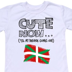 Könnte beinhalten: Weißes T-Shirt mit schwarzem Text, der "Cute Now... (Til My Basque Comes Out)" lautet, und einer rot-grün-weißen Flagge mit einem weißen Kreuz.