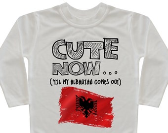 May include: White long-sleeved shirt with the text "CUTE NOW... ('TIL MY ALBANIAN COMES OUT)" in black. Below the text is a red flag with a black double-headed eagle, the flag of Albania.