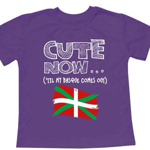 Può includere: Maglietta viola con testo bianco che recita "Cute Now... (Til My Basque Comes Out)" e un contorno bianco della bandiera basca.