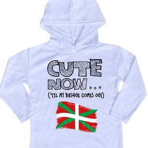 Può includere: Maglietta bianca a maniche lunghe con cappuccio con il testo "Cute Now... (Til my Basque comes out)" e un disegno della bandiera basca.