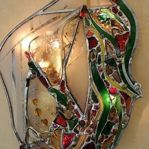 May include: A stained glass wall sconce with a unique abstract design. The sconce features a variety of colours, including red, green, yellow, and gold. The glass is cut into small pieces and arranged in a mosaic pattern. The sconce is illuminated by a light bulb inside.