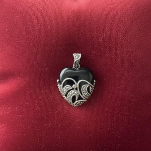Op de afbeelding: Een hartvormige hanger met een zwart stenen midden en sierlijke zilverkleurige filigraan details. De hanger hangt aan een bijpassende balein en rust op een rode ondergrond. Het sieradenstuk is waarschijnlijk een kettinghanger.