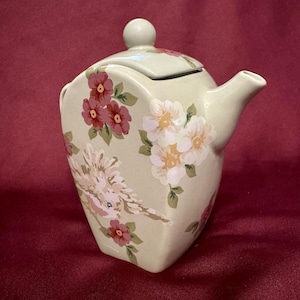 Könnte beinhalten: Eine cremefarbene Keramik-Teekanne mit Deckel und Ausguss. Die Teekanne ist mit rosa und weißen Blumenmustern und einem Vogeldesign verziert. Die Teekanne hat eine abgerundete Form und einen kleinen Griff auf dem Deckel.