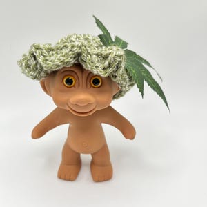 May include: A vintage troll doll with a tan skin tone, large yellow eyes, and a smiling expression. The troll is wearing a green and white crocheted hat with a green leaf accent. The doll is standing against a white background.