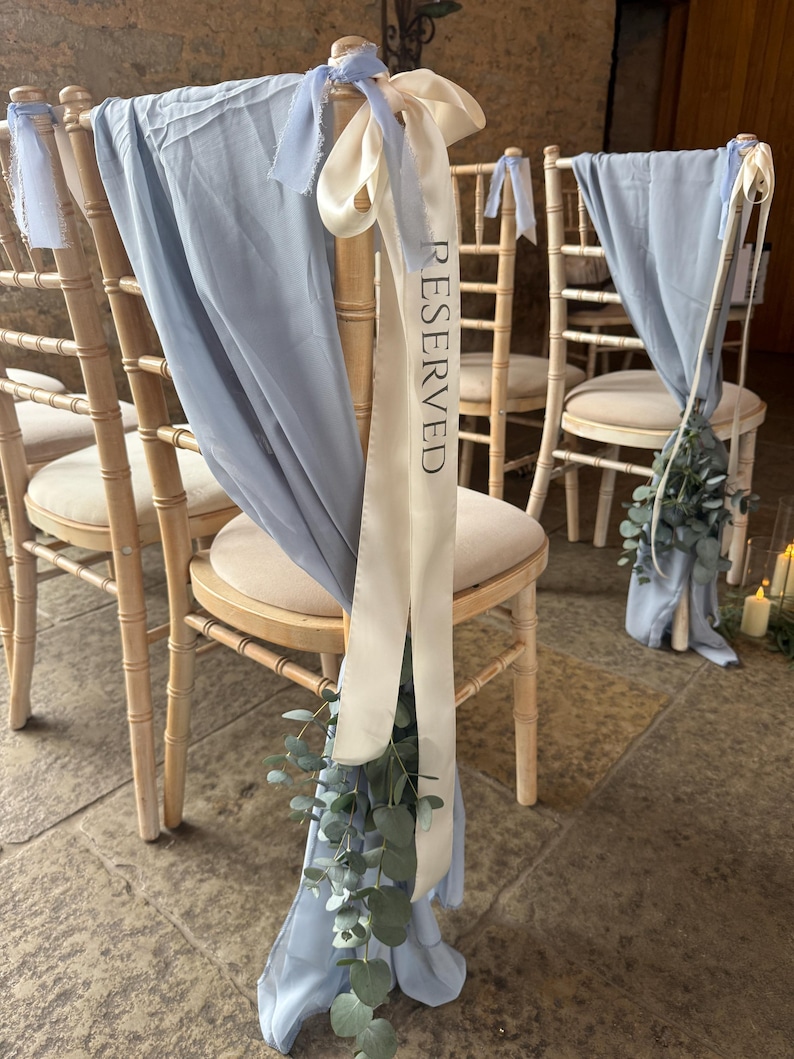 Puede incluir: Sillas de madera clara con cojines color crema, cubiertas con tela azul claro fluida y cintas de sat&eacute;n crema. Una cinta tiene la palabra RESERVED impresa en negro. Cada silla est&aacute; decorada con una peque&ntilde;a rama de hojas de eucalipto verdes.
