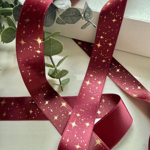 May include: Deep red satin ribbon featuring a gold star design. The ribbon is arranged in loops, with a sprig of greenery in the background. Ideal for gift wrapping and craft projects, this ribbon adds a touch of elegance.