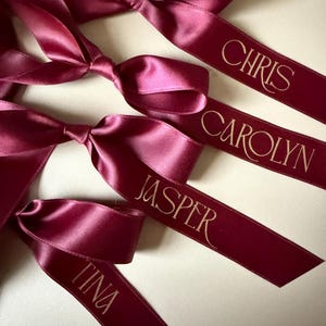 May include: Four deep burgundy satin ribbons tied into bows, each featuring a personalised name printed in a gold serif font. The names displayed on the ribbons are CHRIS, CAROLYN, JASPER, and TINA, arranged in a vertical sequence.