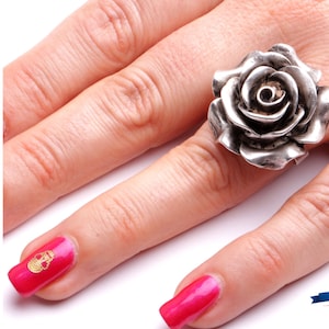 Peut inclure: Une bague en argent avec un motif de rose détaillé. La bague est portée sur un doigt avec un vernis à ongles rose.