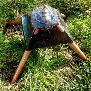 May include: A black pyramid with a copper-colored top and three copper-colored legs. The pyramid is sitting on a bed of green grass.