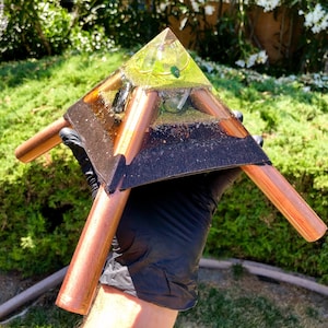 May include: A copper pyramid with a clear resin top containing a green stone. The pyramid is sitting on a black base and has three copper legs.