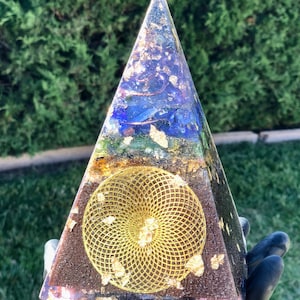 Op de afbeelding: Een heldere glazen piramide met een gouden geometrisch ontwerp erin. De piramide is gelaagd met verschillende gekleurde kristallen en heeft een koperen basis.