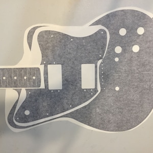 May include: A white guitar body template with a black outline. The template is for a solid body electric guitar with a double cutaway design. The template includes the outline of the body, the neck, the pickup cavities, and the control knobs.