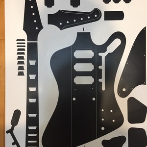 Puede incluir: Una hoja de pegatinas de vinilo negro con una representación gráfica plana y bidimensional de un cuerpo de guitarra eléctrica, un mástil largo con clavijero, varias cubiertas de pastillas, un golpeador y pequeños componentes sobre fondo blanco.