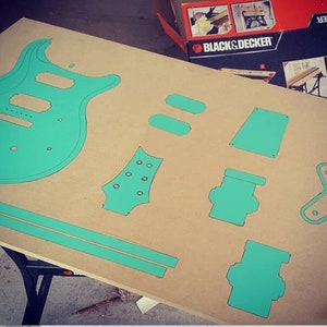 May include: A teal blue guitar body template and other smaller templates for guitar parts are laid out on a brown surface. The templates are cut out of a thin material, possibly cardboard or plywood.