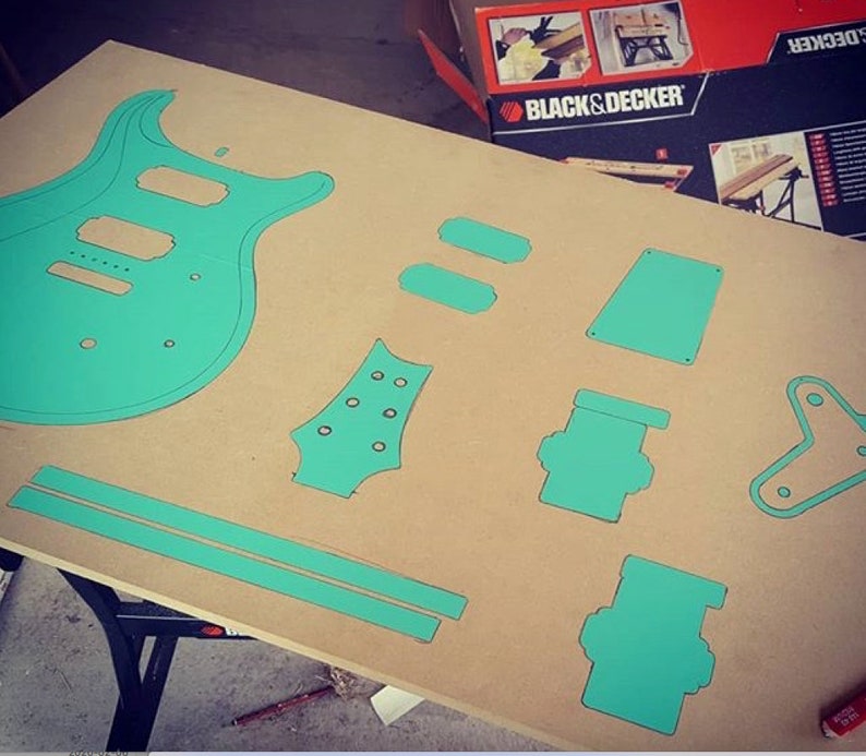 May include: A teal blue guitar body template and other smaller templates for guitar parts are laid out on a brown surface. The templates are cut out of a thin material, possibly cardboard or plywood.