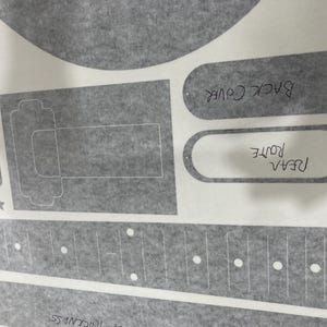 Puede incluir: Una hoja gris con varias plantillas impresas para piezas de guitarra, incluyendo un diapas&oacute;n con marcadores de puntos blancos, una secci&oacute;n de cuerpo rectangular y formas ovaladas etiquetadas "BACK COVER" y "REAR ROUTE" en tinta negra, con cuatro estrellas peque&ntilde;as.