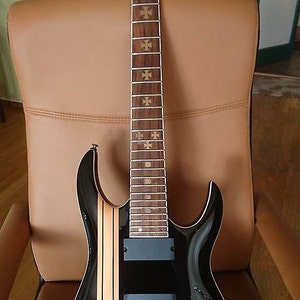 Peut inclure: Guitare électrique noire avec une touche en bois brun et des incrustations de croix dorées. La guitare a un corps noir avec une bande crème au centre. La guitare est fabriquée par la marque "Ibanez".
