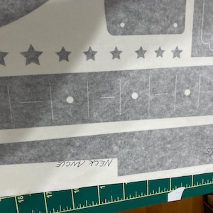 Puede incluir: Una hoja adhesiva gris con varias formas troqueladas, incluyendo una fila de estrellas, tiras rectangulares y patrones geom&eacute;tricos. La hoja incluye etiquetas escritas a mano que dicen NECK ANGLE y THICKNESS sobre una regla verde.