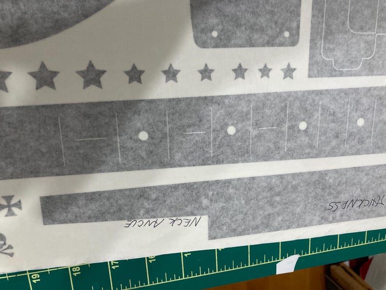 Puede incluir: Una hoja adhesiva gris con varias formas troqueladas, incluyendo una fila de estrellas, tiras rectangulares y patrones geom&eacute;tricos. La hoja incluye etiquetas escritas a mano que dicen NECK ANGLE y THICKNESS sobre una regla verde.