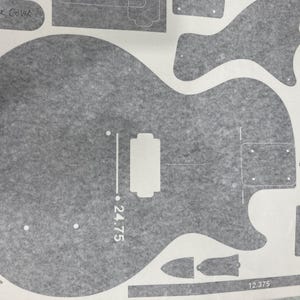 Puede incluir: Una hoja de plantilla gris con varias piezas de guitarra, incluyendo el contorno de un cuerpo de guitarra con una marca de escala de 62,865 cm, un clavijero, cavidades para pastillas y una tapa trasera sobre un fondo gris texturizado.