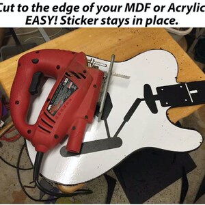 May include: A red electric jigsaw tool with a black cord is being used to cut a white guitar shape with a black sticker on it. The text "Cut to the edge of your MDF or Acrylic. EASY! Sticker stays in place." is visible at the top of the image.