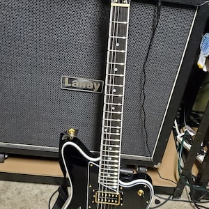 customer build surfcaster Black electric guitar with gold hardware and a white pickguard. The guitar is leaning against a black amplifier with a 'Laney' logo.