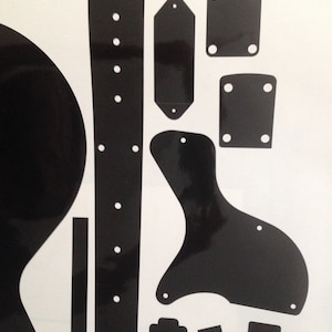 Puede incluir: Un conjunto de componentes de guitarra negros, brillantes y planos dispuestos sobre una superficie blanca. El set incluye una tira larga y delgada con orificios circulares, un golpeador curvo, placas rectangulares con orificios y otras formas.