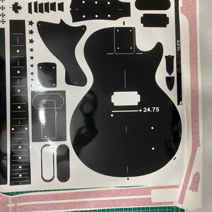 Puede incluir: Una hoja de pegatinas de vinilo negro con varios componentes de guitarra, incluyendo el contorno del cuerpo, el diapas&oacute;n, el clavijero y formas de herrajes. El dise&ntilde;o incluye el texto 24.75 y 12.375, junto con iconos de estrellas y hojas de arce.