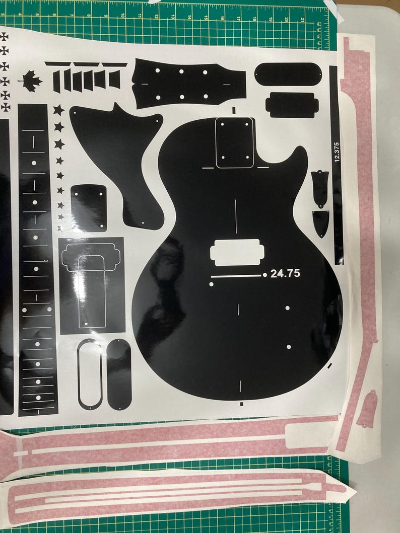 Puede incluir: Una hoja de pegatinas de vinilo negro con varios componentes de guitarra, incluyendo el contorno del cuerpo, el diapas&oacute;n, el clavijero y formas de herrajes. El dise&ntilde;o incluye el texto 24.75 y 12.375, junto con iconos de estrellas y hojas de arce.