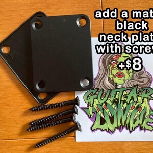 May include: Two black metal neck plates with screws for a guitar. The plates are on a wooden surface. A sticker with a zombie hand and the text "Guitar Zombie" is in the background. The text "add a matte black neck plate with screws +&pound;8" is also visible.